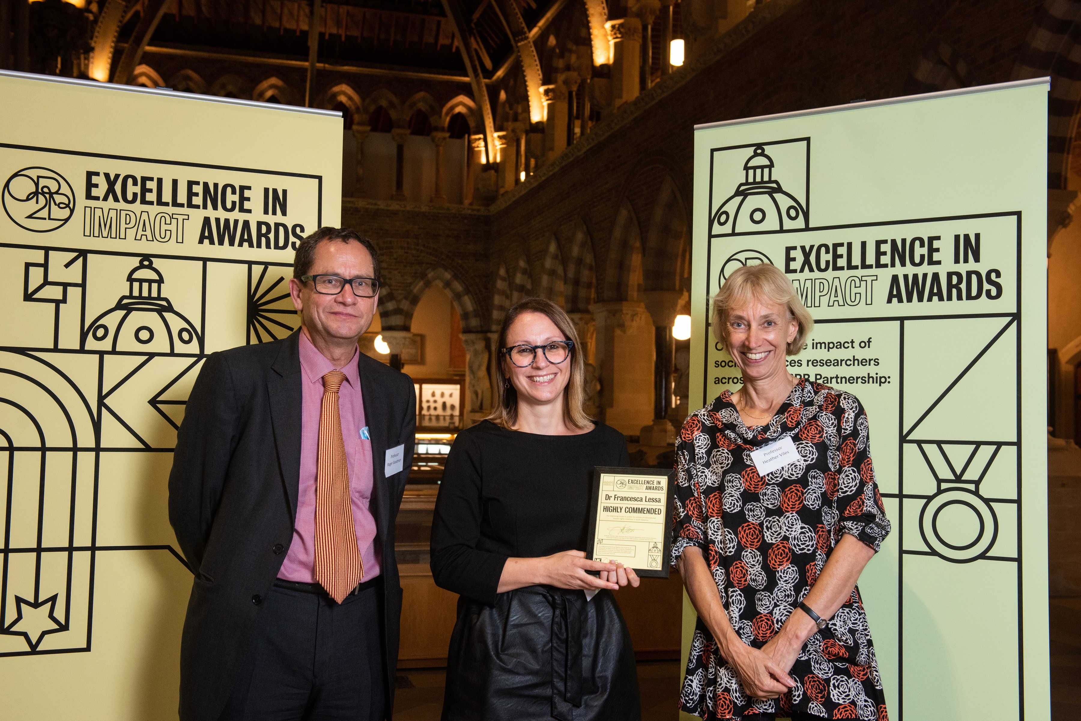 Dr Francesca Lessa holds her Excellence in Impact Awards 'Highly Commended' certificate in front of the event branding, flanked by Professor Roger Goodman and Prof Heather Viles 