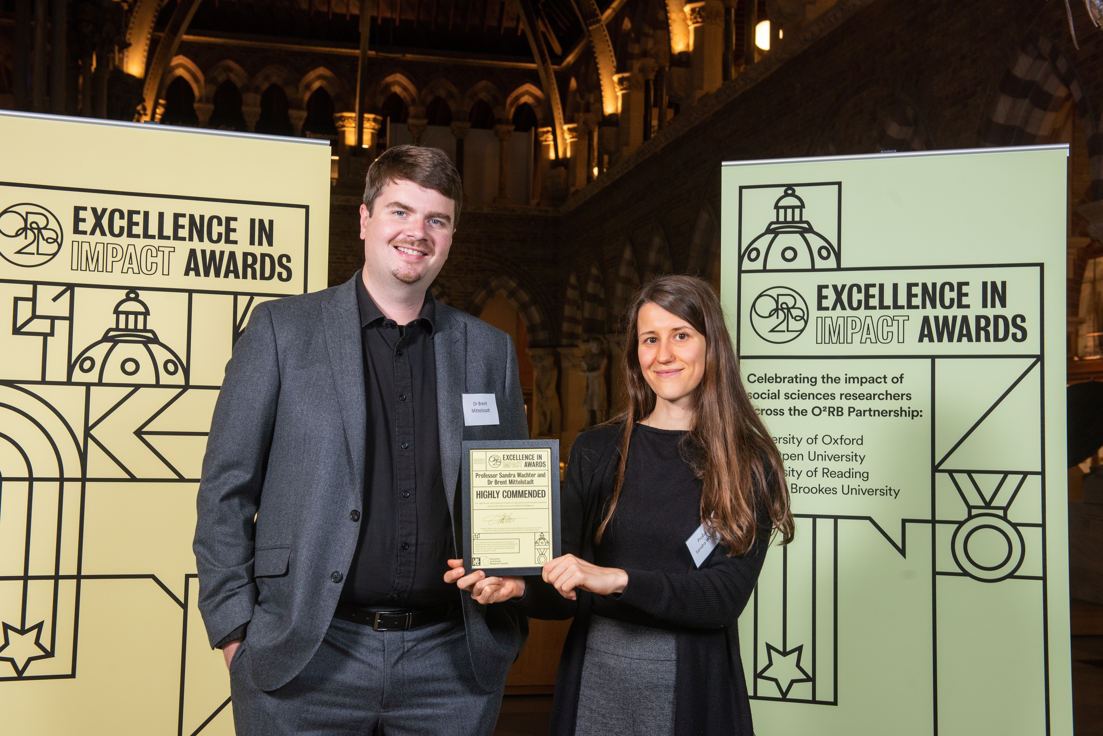 Professor Sandra Wachter and Dr Brent Mittelstadt hold their Highly Commended impact award, flanked by event branding at the awards reception