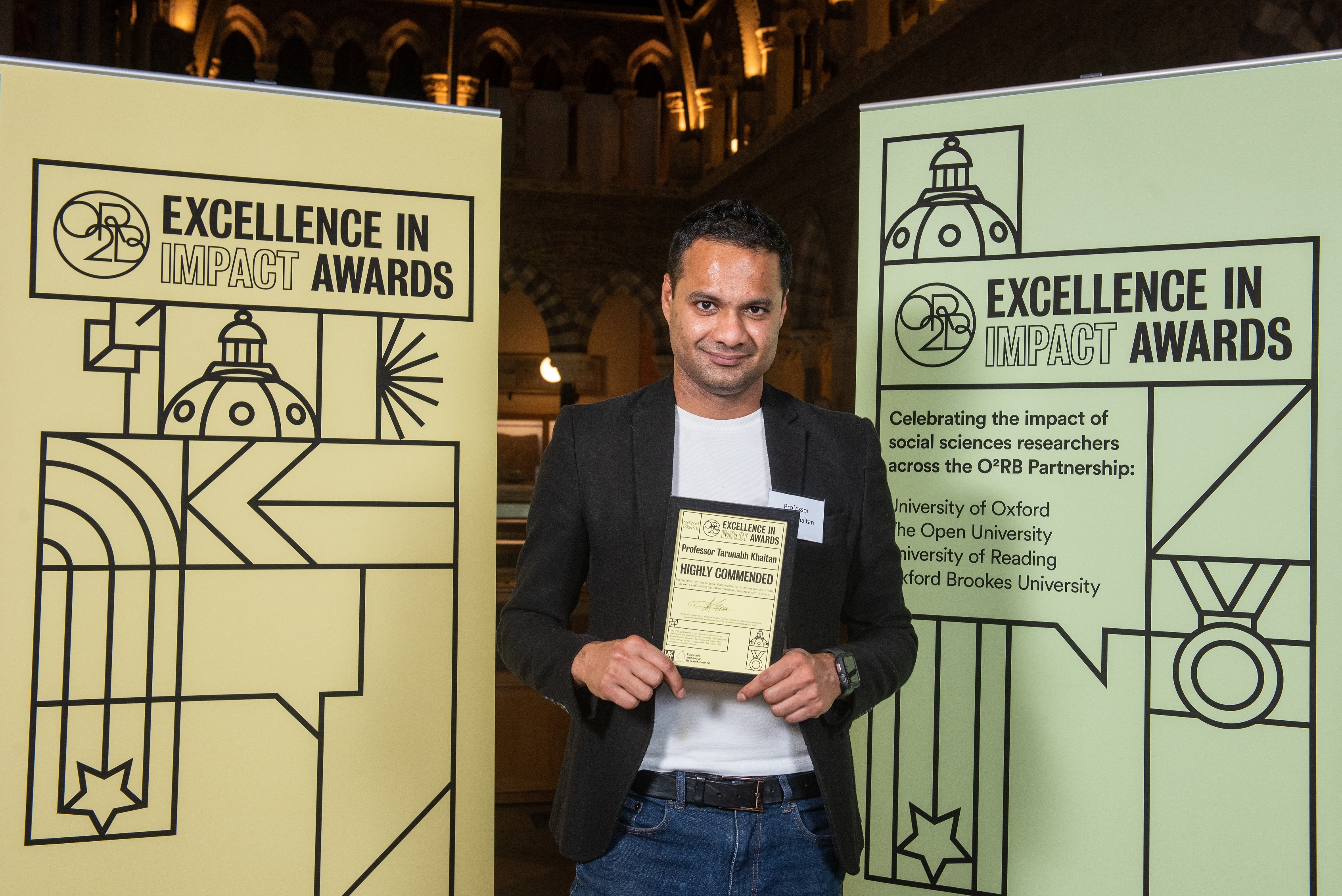 Professor Tarunabh Khaitan holds his Highly Commended impact award, flanked by event branding at the awards reception
