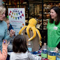 Children and adult at welcome desk to the Festival of Social Science, talking to event lead. Table has maps and leaflets on, and a feedback box with a yellow toy octopus on top. 