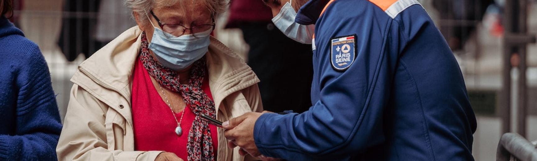 A woman wearing a mask in a queue of people presents vaccine documentation to an official permitting entry