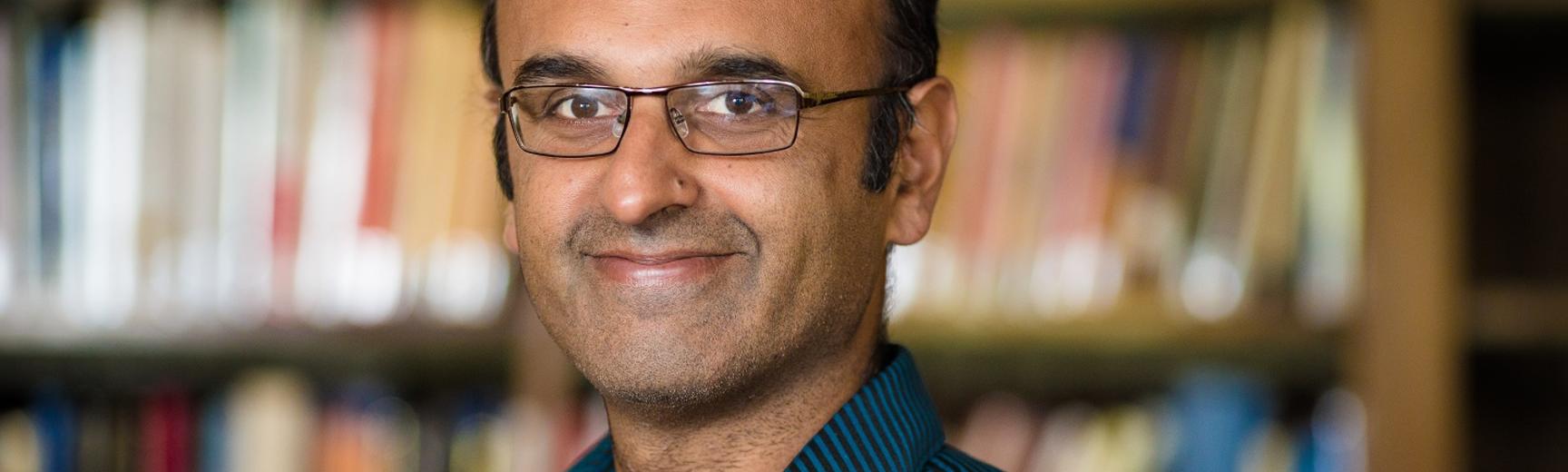 Portrait photo of Professor Yadvinder Malhi, smiling against a blurred background of shelves full of books
