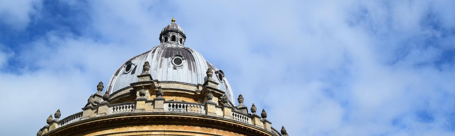 A view of the circular Radcliffe Camera library showing the blue-grey leaded roof and yellow standstone walls against a bright blue sky with whispy clouds