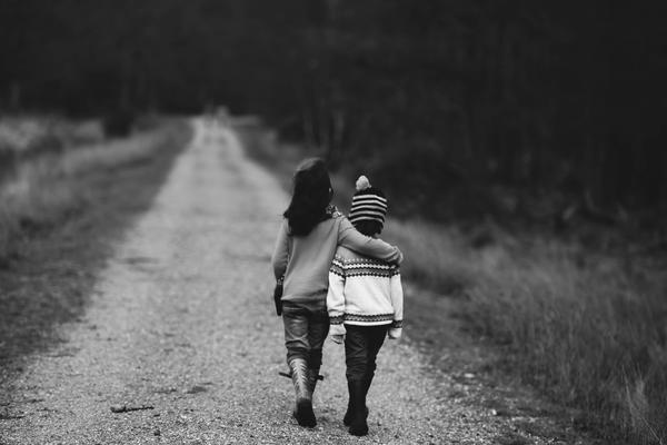 A black and white photo of two small children walking down a track, away from the camera. The bigger child has their arm around the smaller one, as if comforting them.