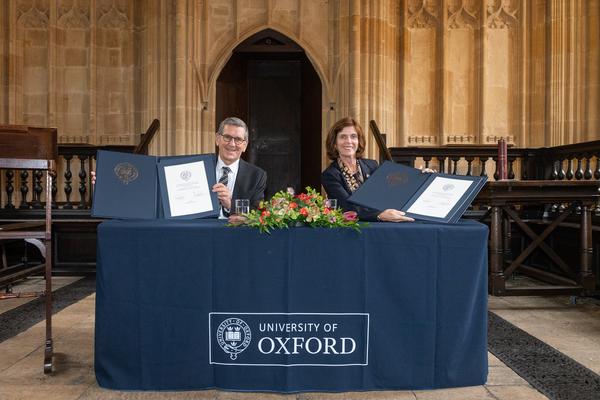 Professor Louise Richardson (VC of University of Oxford) and Professor Reinhold Geilsdoerfer (MD of the Dieter Schwarz Foundation) sit at an Oxford-blue branded desk hoding signed agreements and smiling