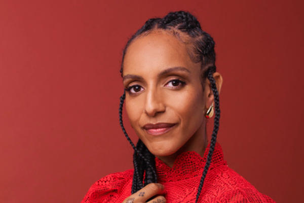 Portrait of Afua Hirsch, a Black woman with long braids, wearing a red lace top and gold jewellery, posing confidently against a warm red background. Her hand, adorned with a dotted design, rests over her chest.