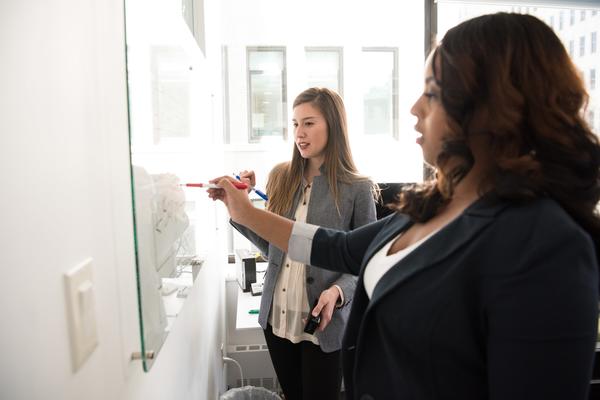 Two women of diverse ethnicities work together on a whiteboard using different coloured pens