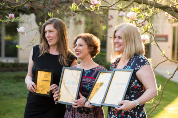 Three researchers stand in a sunny garden with a lawn and trees behind them, holding their prize certificates and smiling for the camera at the Excellence in Impact Awards