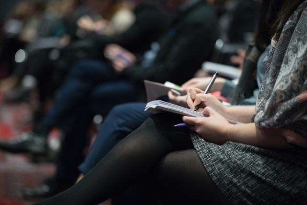 A row of people seated at an event, showing only their legs and the laps. In the forground, a person wearng a grey dress is taking notes on a notepad on her lap.