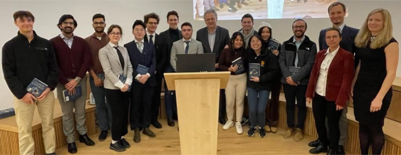 Seventeen smiling, smartly dressed people stand behind a lectern, some holding copies of a book. 