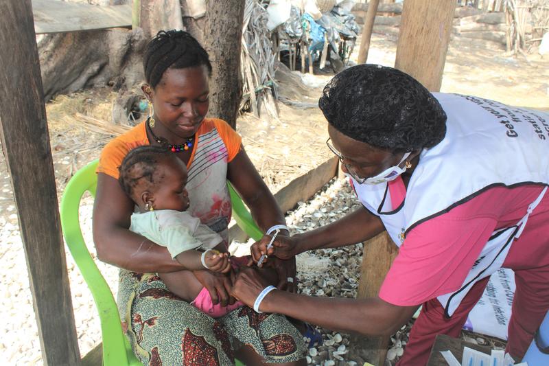 A young Sierra Leonean woman sits in an open-air clinic with a baby on her lap, whilst a female nurse administers an injection into the baby's leg. 