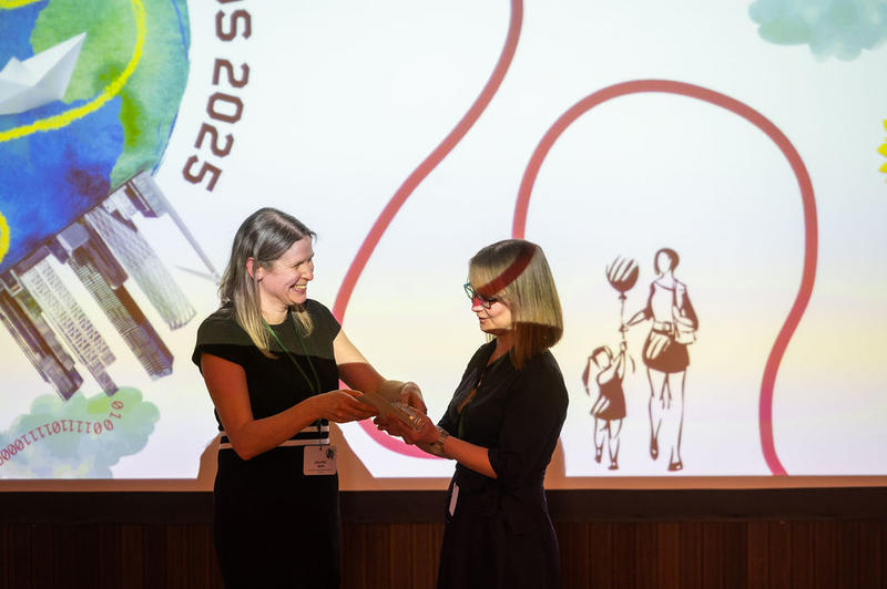Dr Jen Gold presents the Emerging Impact trophy to Dr Jennifer Seddon Dr Jen Gold smiles as she hands over the Emerging Impact Award trophy to Dr Jennifer Seddon in front of a screen with award branding