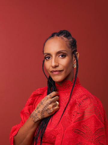 Portrait of Afua Hirsch, a Black woman with long braids, wearing a red lace top and gold jewellery, posing confidently against a warm red background. Her hand, adorned with a dotted design, rests over her chest.