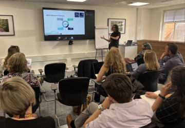 A room of seated people listening to Prof Rebecca Williams present the AIEOU Hub