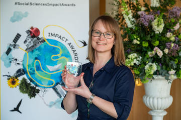 Dr Jennifer Seddon smiles and holds her prize trophy. In the background is the Impact Awards branding and a flower arrangement.