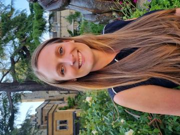 Naomi is standing in a garden smiling at the camera. She has long blonde hair and is wearing a navy blouse.