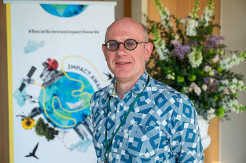  Dr Michael Biggs smiles to camera. In the background is the Impact Awards branding and a flower arrangement.