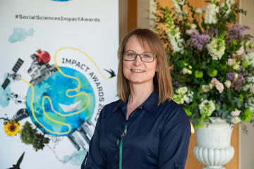 Dr Jennifer Seddon smiles to camera. In the background is the Impact Awards branding and a flower arrangement.