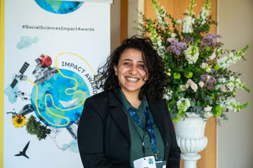 Dr Mona Ibrahim smiles to camera. In the background is the Impact Awards branding and a flower arrangement.