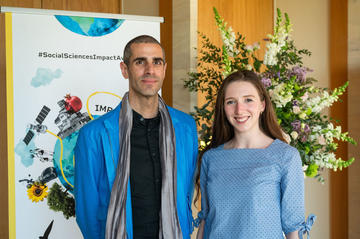 Dr Six Silberman and Aislinn Kelly-Lyth smile to camera.  In the background is the Impact Awards branding and a flower arrangement.