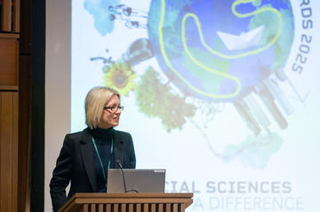 Prof Louise Slater Impact Awards Prof Louise Slater speaks from a lectern in front of a screen with the event branding