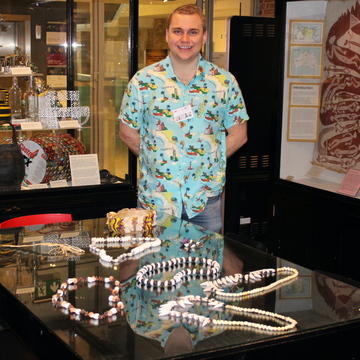 Dr Liam Saddington, wearing a colourful island-themed tropical shirt, stands smiling in front of a table full of shell necklaces and paraphernalia from islands in the South Pacific