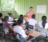 Professor Laura Rival smiles as she stands at the front of classroom, teaching a class of young adults. The classroom walls are bright green, the students and teacher are wearing clothes for hot weather, and out of the window a lush rainforest is visible.