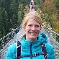 Dr Amy Styring Dr Styring is standing on a suspension bridge, smiling. She has blonde hair which is tied back, and is wearing a bright blue coat with a black rucksack.