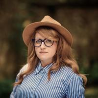 Dr Gemma Newlands Dr Gemma Newlands has long auburn wavy hair and large, blue-rimmed glasses. She is wearing a straw hat and a blue and white striped shirt, looking directly at the camera.